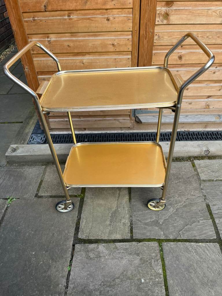 A gold coloured metal tea trolley on wheels and with two serving trays.