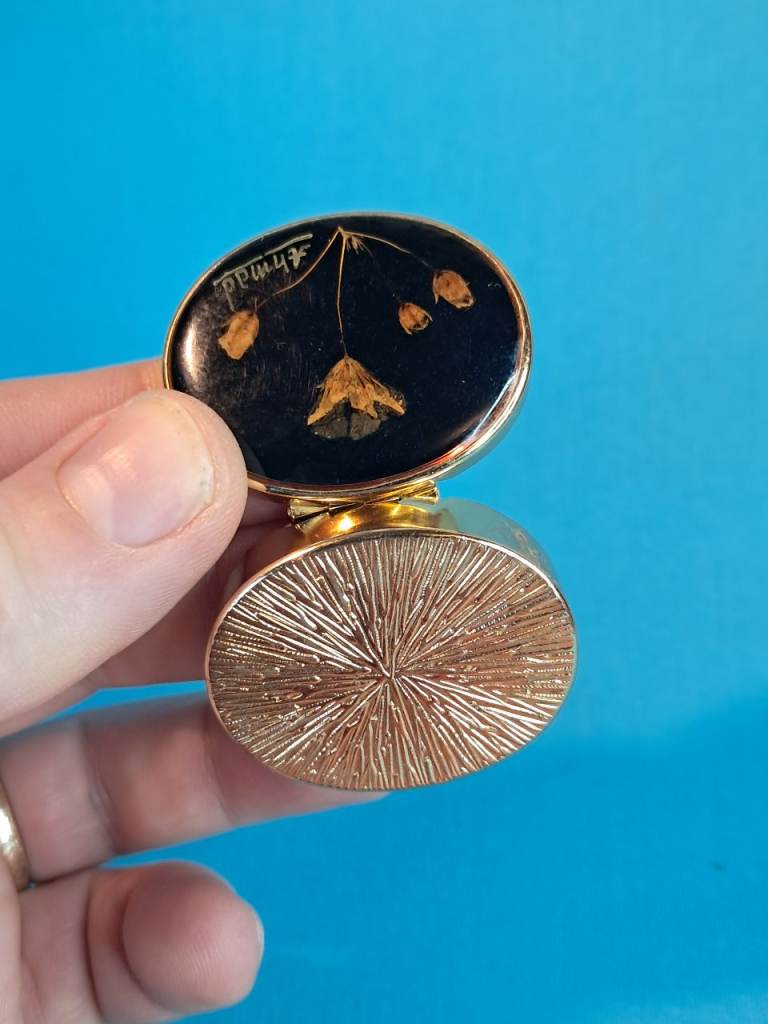 gold-tone metal pill box with a black resin top featuring real pressed flowers and the name 'Ahmad.